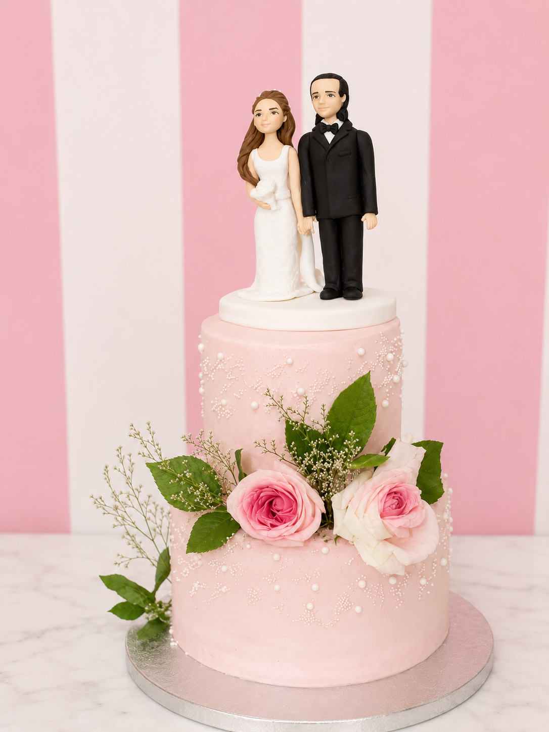 A two-tier pink wedding cake with white frosting, decorated with pink and white roses, green leaves, and white pearl-like accents. A custom bride and groom figurine stands on top of the cake, set against a pink and white striped background.
