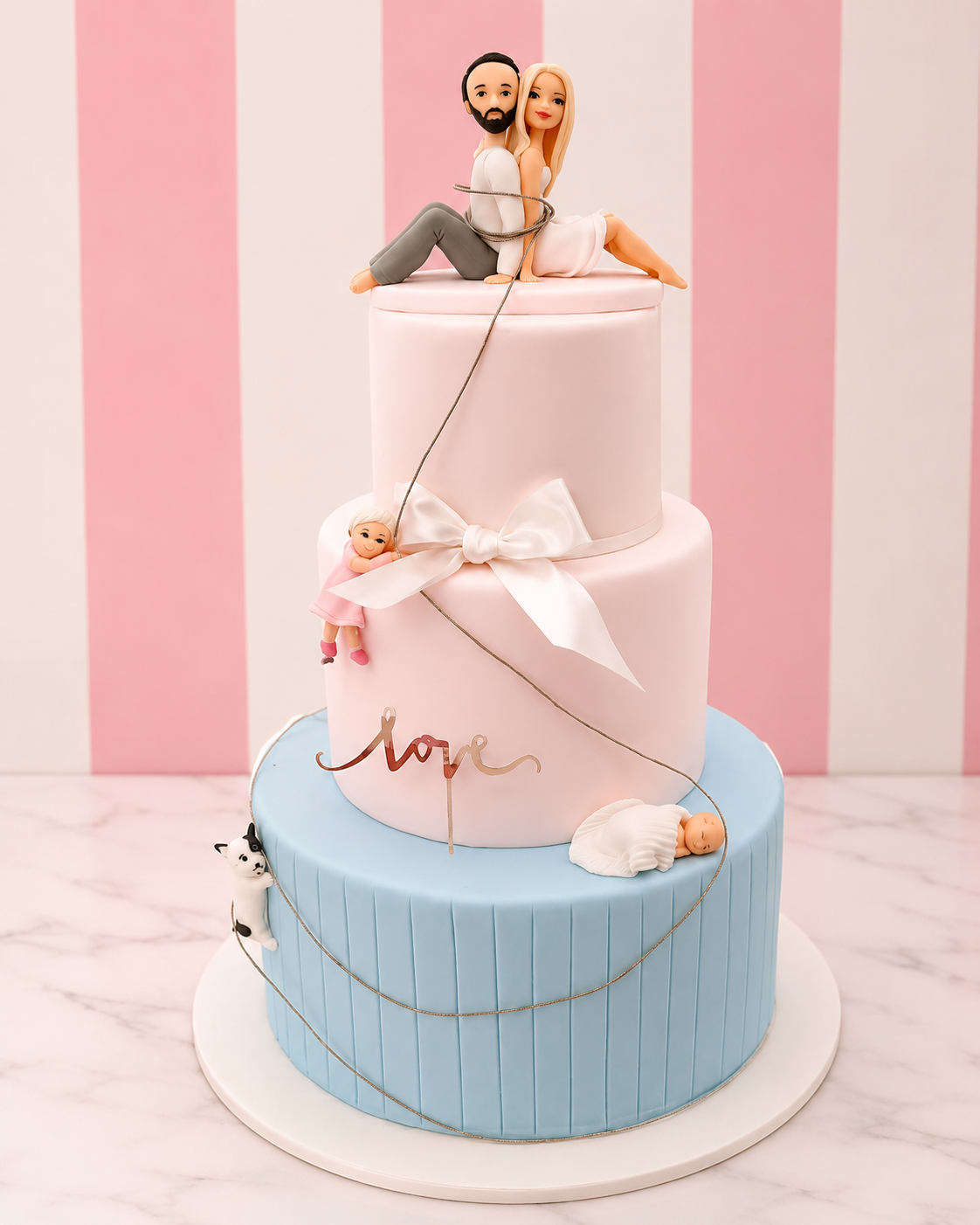A three-tier custom fondant cake with a pink and light blue color scheme, decorated with a white bow, gold script 'love' lettering, and baby figurines, topped with a couple's figurine.